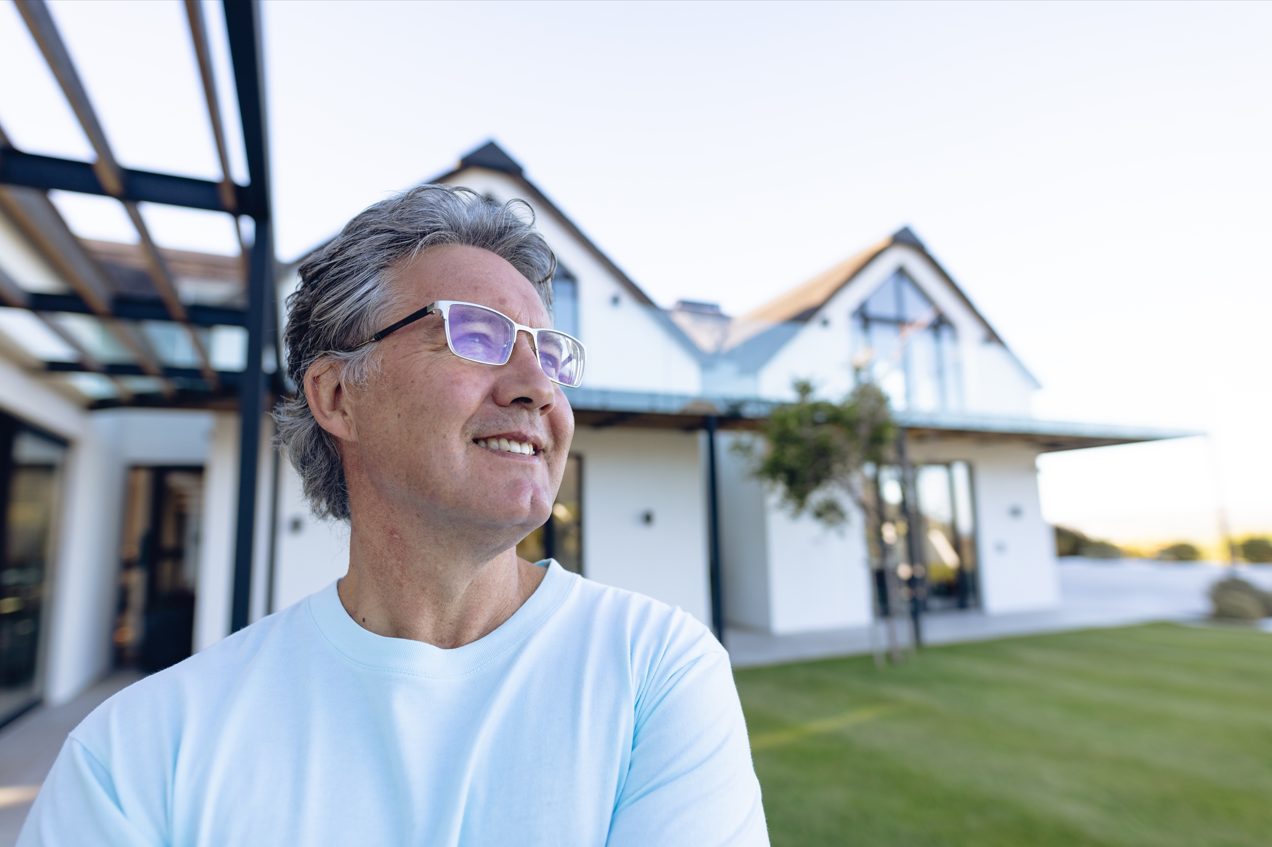 Smiling caucasian senior man wearing eyeglasses looking away against house in yard after learning about types of mortgages that would help him achieve homeownership.